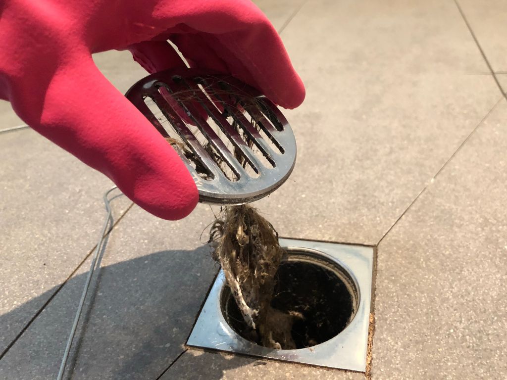 Gloved hand removing a hair clog from a shower drain, showing a common cause of clogged drains and slow water drainage.
